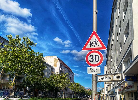 Tempo 30 Anordnung auf einer Berliner Hauptverkehrsstraße: Hier ist ein Schulweg. Auf dem Foto sind zwei Verkehrsschilder vor blauem Himmel zu sehen: Tempo 30 und Kinder. Rechts liegt eine Häuserzeile, auf der linken Seite ist die Straße durch Bäume begrünt.