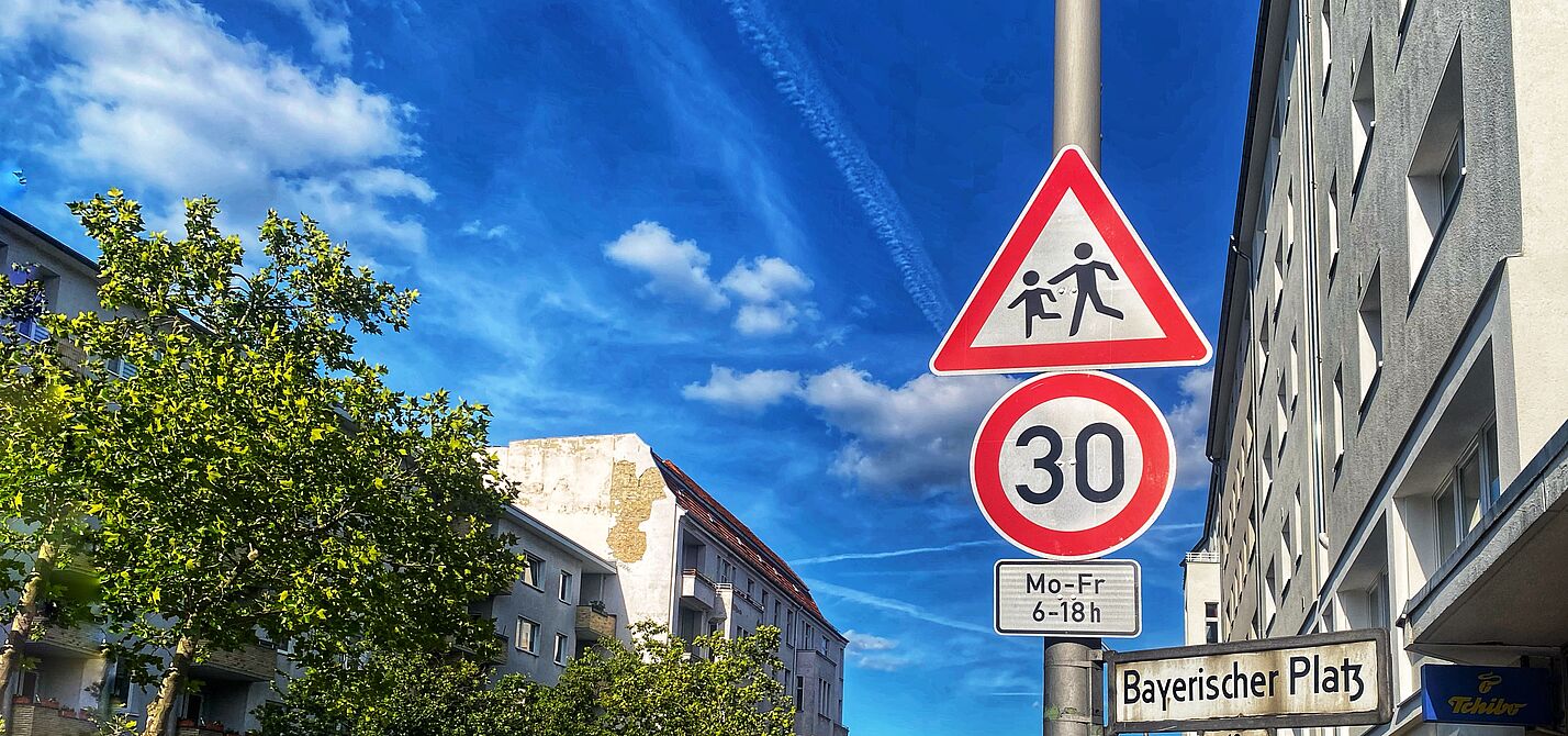 Tempo 30 Anordnung auf einer Berliner Hauptverkehrsstraße: Hier ist ein Schulweg. Auf dem Foto sind zwei Verkehrsschilder vor blauem Himmel zu sehen: Tempo 30 und Kinder. Rechts liegt eine Häuserzeile, auf der linken Seite ist die Straße durch Bäume begrünt.