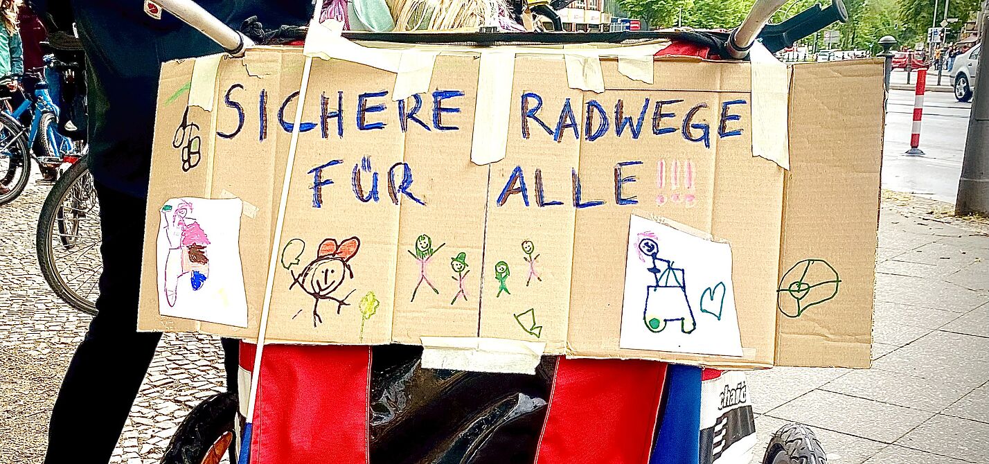 Sichere Radwege für alle! Auf dem Foto ist ein Pappschild zu seinen, dass an der Rückseite eines Fahrradanhängers angebracht ist. Neben bunten Kinderzeichnungen ist in gemalten Großbuchstaben „sichere Radwege für alle“ zu lesen.