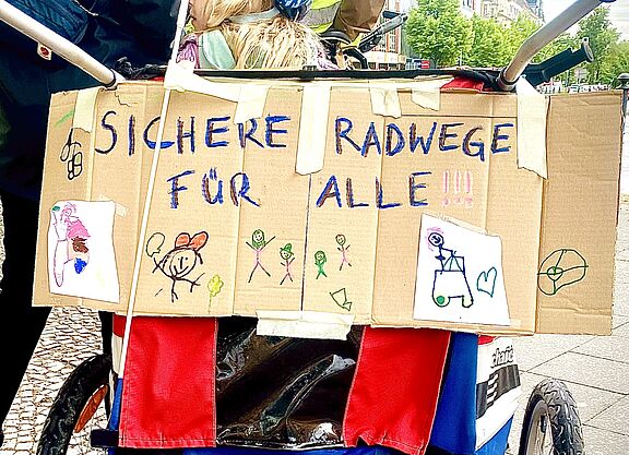 Sichere Radwege für alle! Auf dem Foto ist ein Pappschild zu seinen, dass an der Rückseite eines Fahrradanhängers angebracht ist. Neben bunten Kinderzeichnungen ist in gemalten Großbuchstaben „sichere Radwege für alle“ zu lesen.