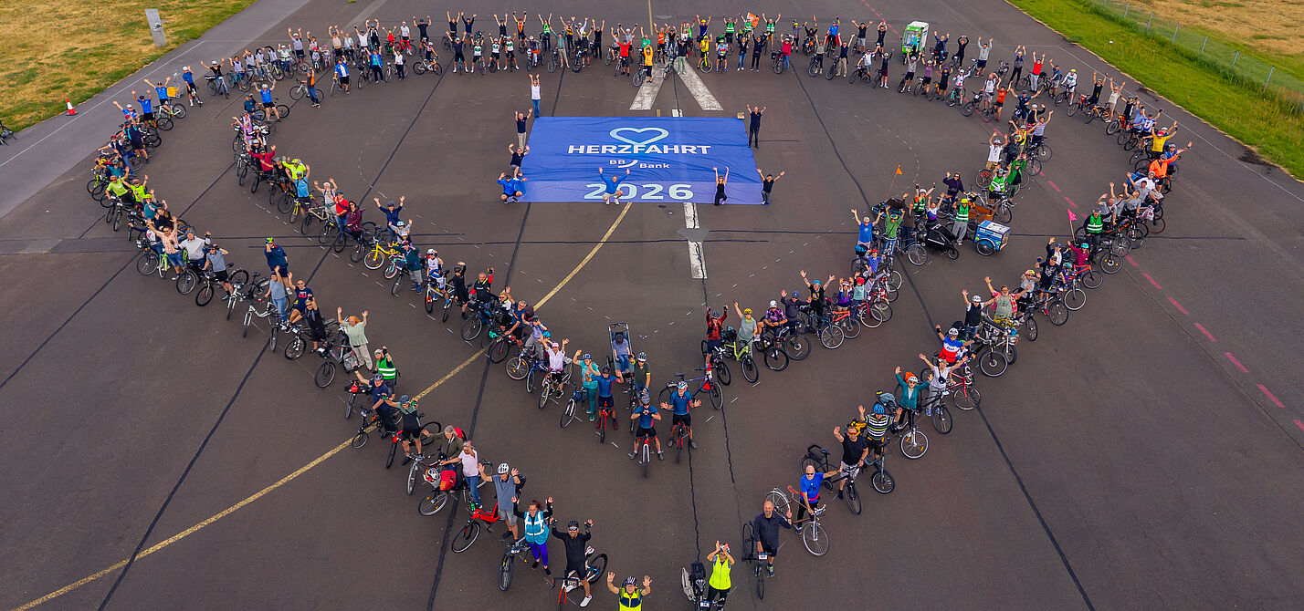 HerzFahrt 2026 Menschen mit Fahrrädern bilden zwei Herzen um HerzFahrt Banner auf dem Boden