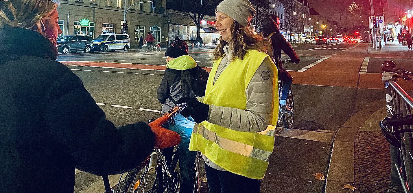 ADFC Lebkuchen-Aktion in Berlin Schöneberg Eine Frau mit gelber Warnweste steht an einer roten Ampel in Berlin Schöneberg, Hauptstraße Ecke Kolonnenstraße, und übergibt einer Radfahrerin eine Karte des ADFC mit einem Lebkuchen.