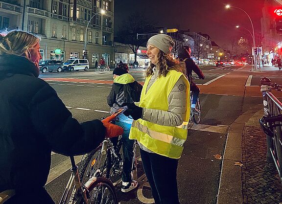 ADFC Lebkuchen-Aktion in Berlin Schöneberg Eine Frau mit gelber Warnweste steht an einer roten Ampel in Berlin Schöneberg, Hauptstraße Ecke Kolonnenstraße, und übergibt einer Radfahrerin eine Karte des ADFC mit einem Lebkuchen.