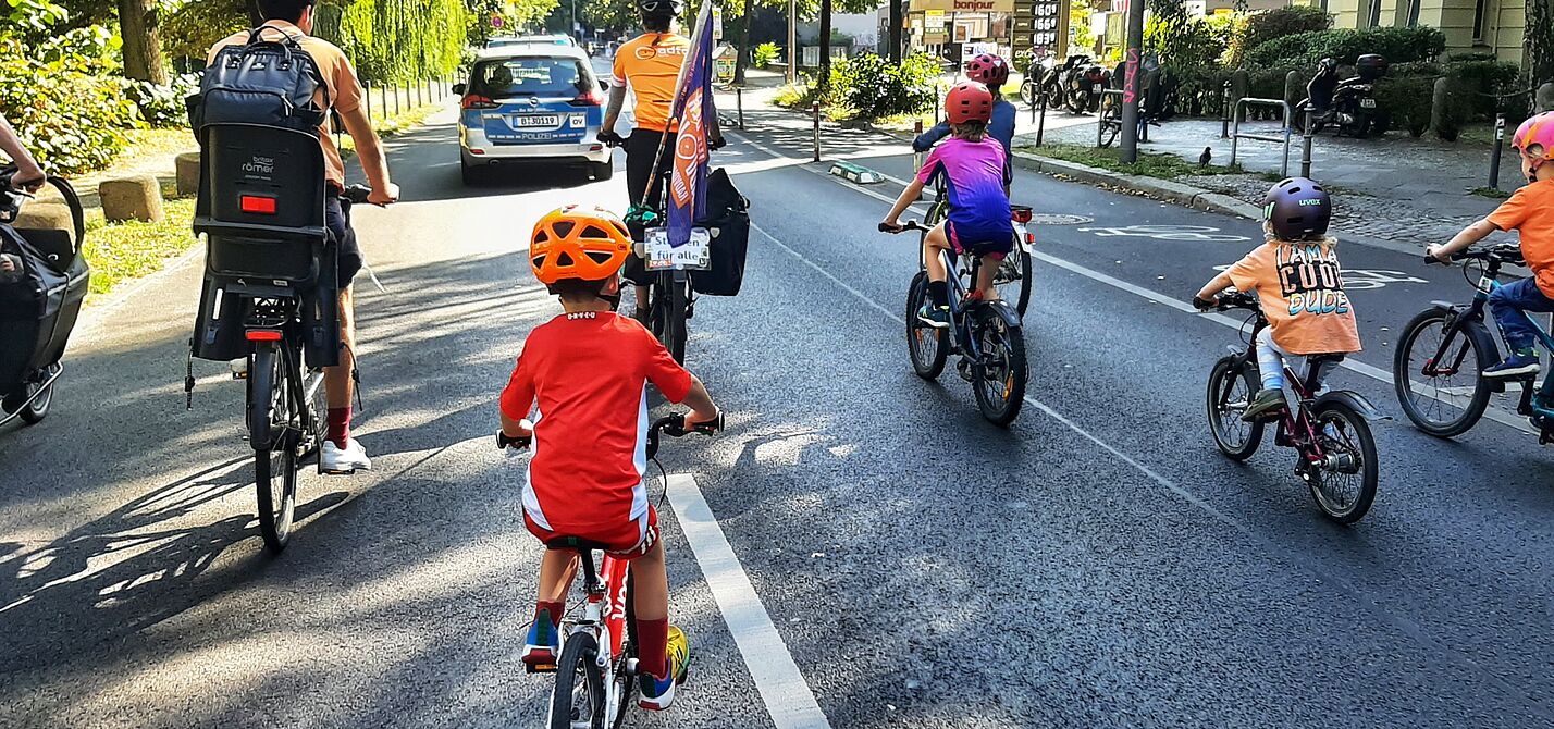 Eine Gruppe Kinder fährt bei sonnigem Wetter auf Fahrrädern das Berliner Reichpietschufer entlang, auf der Fahrbahn. Vorne fährt ein Auto der Polizei Berlin als Führungsfahrzeug, und zwei Erwachsene. Es ist eine ADFC Berlin Fahne zu sehen.