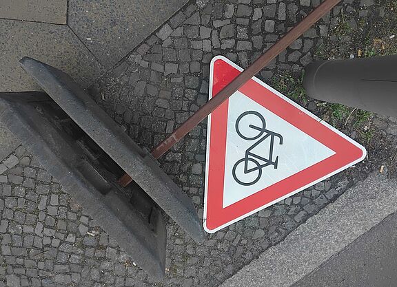 Das Bild zeigt ein rotes dreieckiges "Achtung Radfahrer"-Schild, es liegt auf dem Boden neben dem Fuß eines Baustellen-Aufstellers