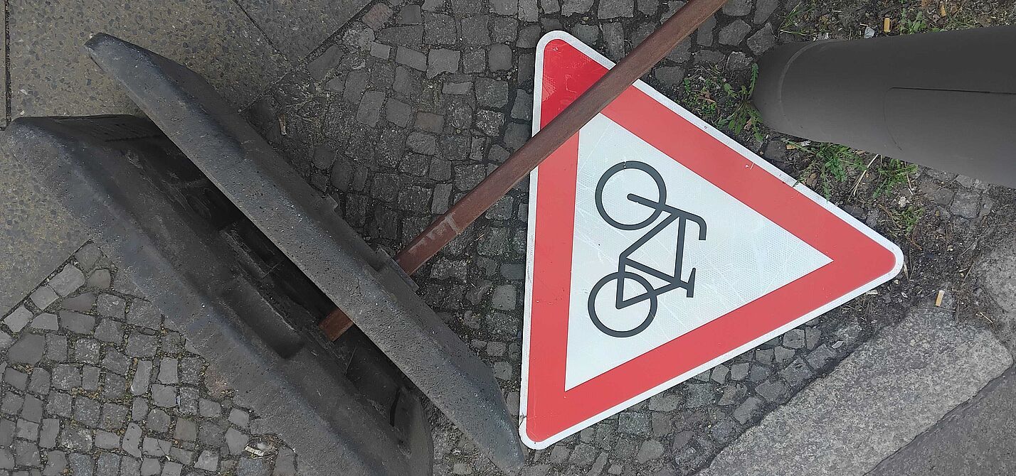 Radwegestopp 2.0? Das Bild zeigt ein rotes dreieckiges "Achtung Radfahrer"-Schild, es liegt auf dem Boden neben dem Fuß eines Baustellen-Aufstellers