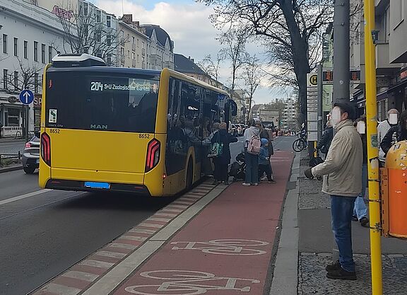 Haltestellenkap in der Hauptstraße in Schöneberg Das Bild zeigt im rechten Bereich eine Haltestelle, daneben ein Haltestellenkap mit darüber geführten Radweg und ein haltender Bus. Fahrgäste steigen ein und aus.