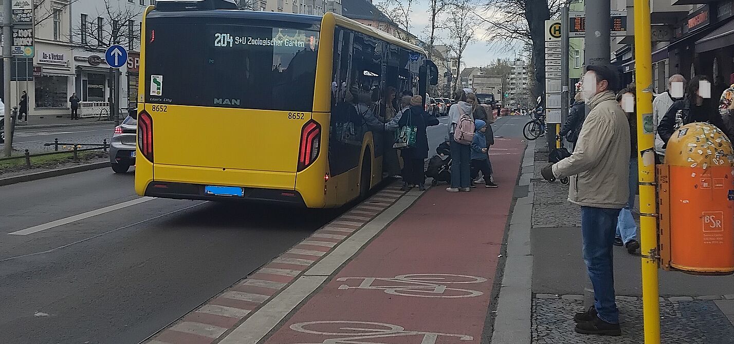 Haltestellenkap in der Hauptstraße in Schöneberg Das Bild zeigt im rechten Bereich eine Haltestelle, daneben ein Haltestellenkap mit darüber geführten Radweg und ein haltender Bus. Fahrgäste steigen ein und aus.