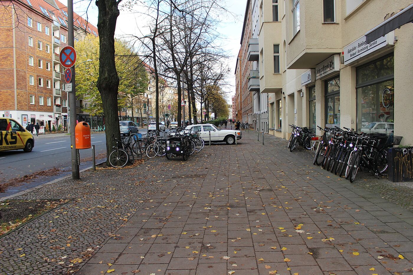 Gehweg breit breiter Gehweg einer Stadtstraße, rechts querstehende Fahrräder vor einem Fahrradgeschäft, links geparkte Fahrräder, ein querstehendes Auto in einer Einfahrt, im Hintergrund Fußgänger:innen