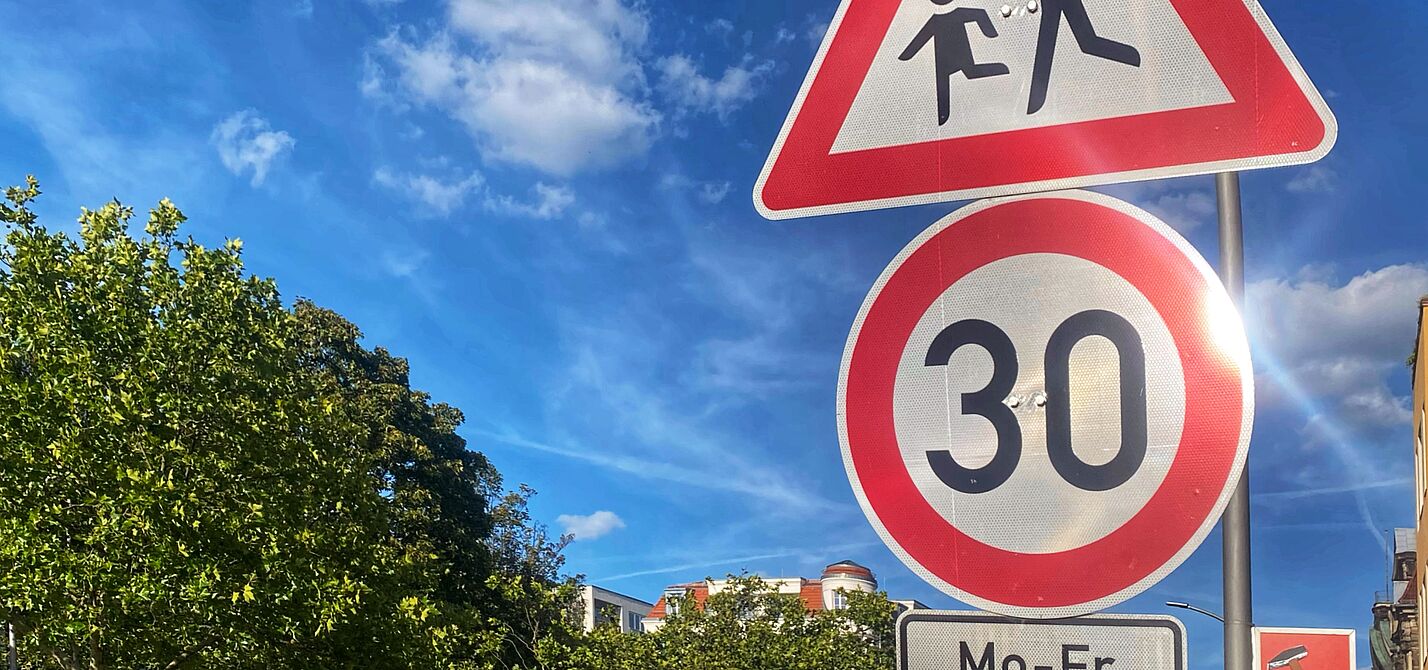 Auf dem Foto sind zwei Verkehrsschilder vor blauem Himmel zu sehen: Tempo 30 und Kinder. Rechts liegt eine Häuserzeile, auf der linken Seite ist die Straße durch Bäume begrünt.