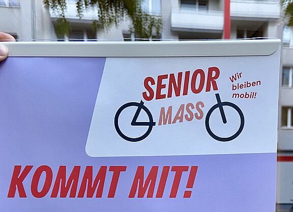 Senior Mass Plakat von Changing Cities Ein Plakat mit dem Titel „Senior Mass – Kommt mit!“ wird vor einer Schaufensterscheibe ins Licht gehalten.