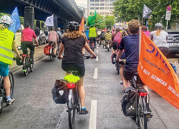 Eine Gruppe Radfahrende fährt mit Fahnen auf einer Fahrbahn Richtung Kreuzberg. Links im Bild ist die oberirdische U-Bahnlinie 1 zu sehen. 