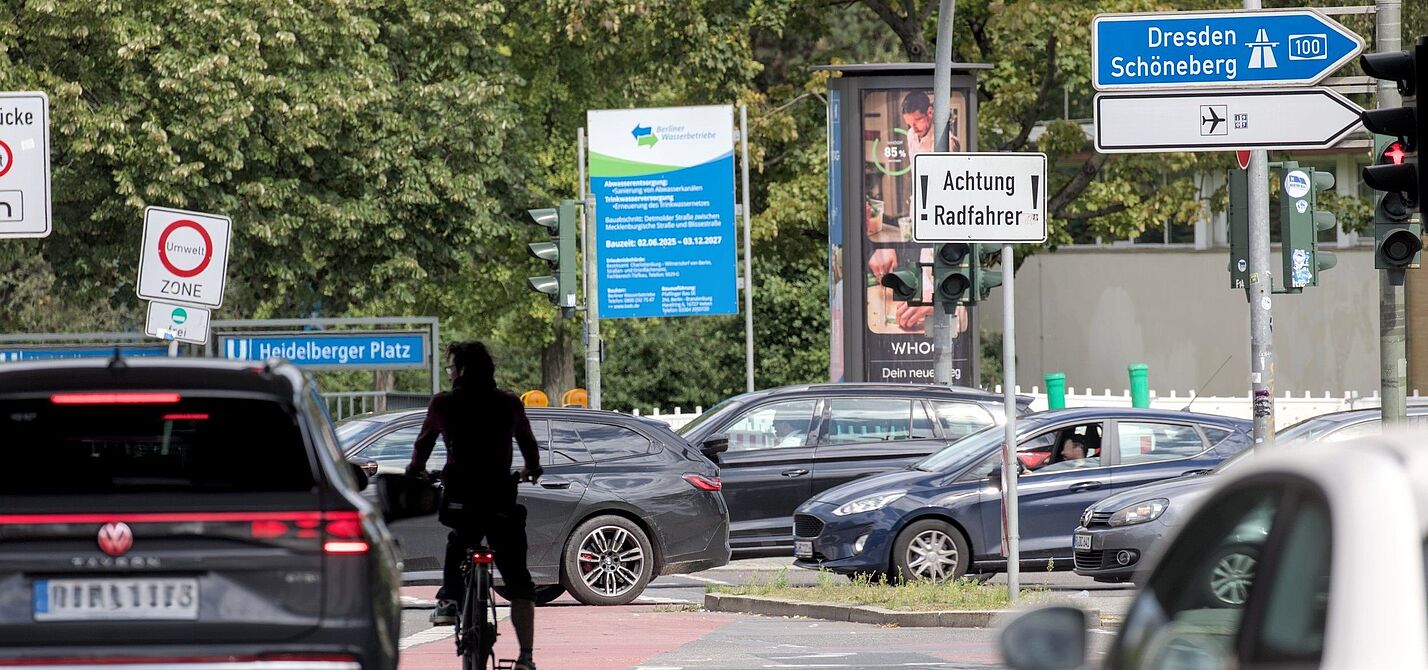 Radfahren in Berlin, Mecklenburgische Straße, Heidelberger Platz Auf einer Hauptverkehrsstraße am Heidelberger Platz in Berlin ist unter einer Brücke nahe einer Auffahrt zur A100 die Silhoutte eines Radfahrers zu sehen. Er steht mit seinem Rad mitten auf der Fahrbahn, umgeben von Autos. Zum Radfahren ist dort wenig Platz. Es sind mehrere Schilder zu sehen, unter anderem ein weißes Schild, das die Aufschrift „Achtung Radfahrer“ trägt.