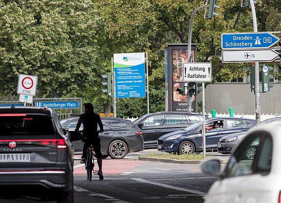 Radfahren in Berlin, Mecklenburgische Straße, Heidelberger Platz Auf einer Hauptverkehrsstraße am Heidelberger Platz in Berlin ist unter einer Brücke nahe einer Auffahrt zur A100 die Silhoutte eines Radfahrers zu sehen. Er steht mit seinem Rad mitten auf der Fahrbahn, umgeben von Autos. Zum Radfahren ist dort wenig Platz. Es sind mehrere Schilder zu sehen, unter anderem ein weißes Schild, das die Aufschrift „Achtung Radfahrer“ trägt.