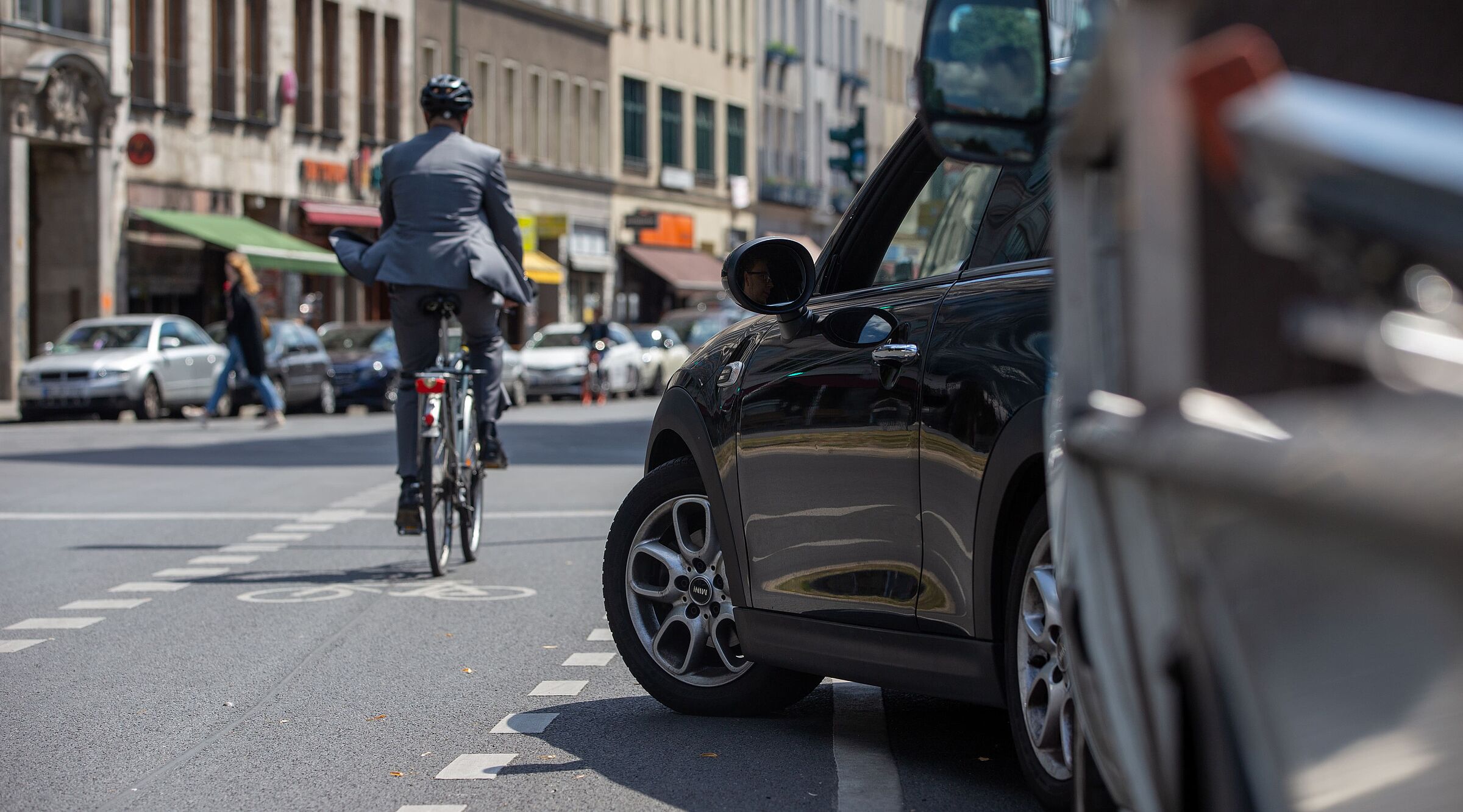 Radfahrer in dunkler Kleidung mit Helm fährt auf einer Stadtstraße, im Vordergrund steht ein schwarzes Auto am Straßenrand, weitere Fahrzeuge und Gebäude im Hintergrund.