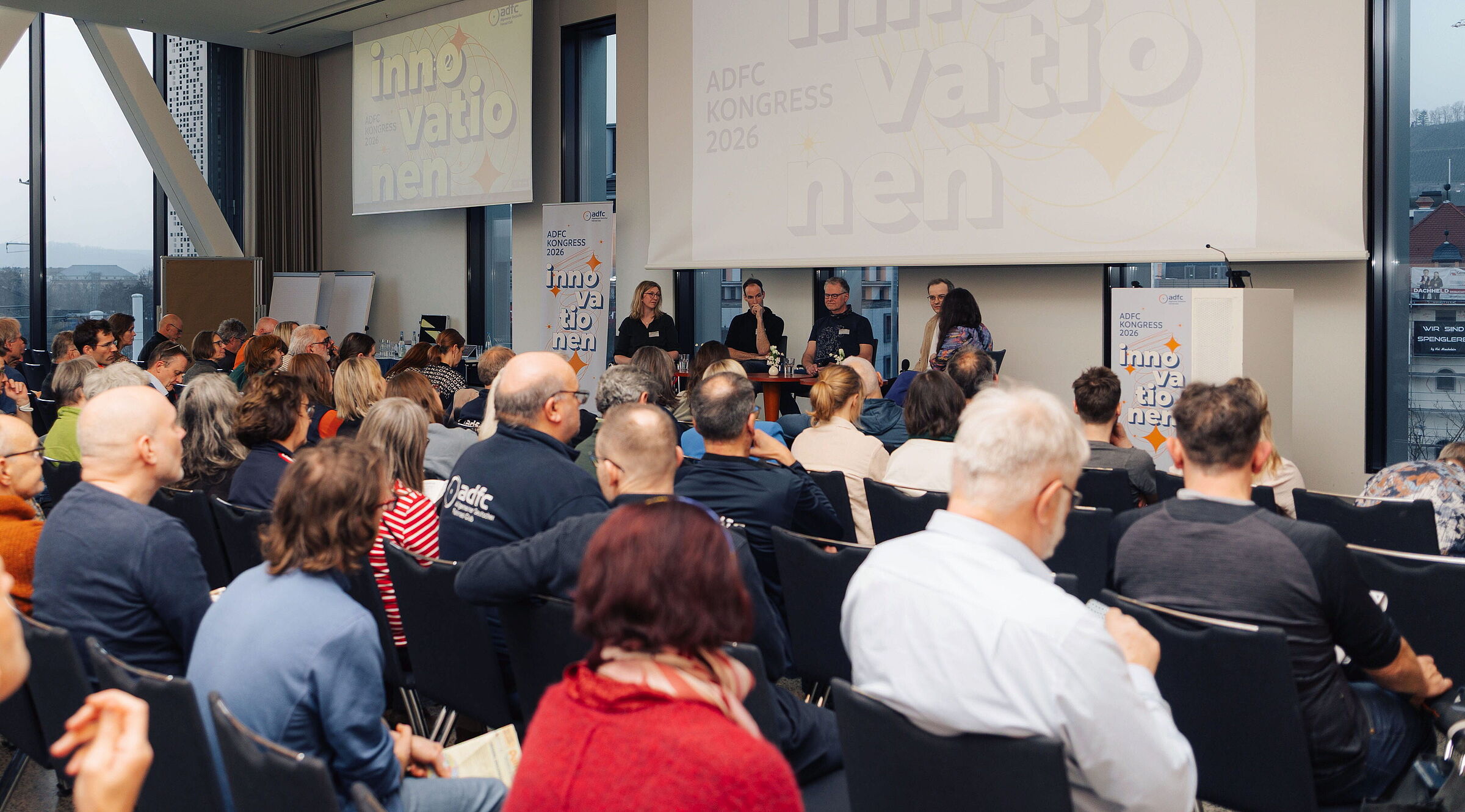 Blick über die Köpfe zahlreicher sitzender Zuschauer im Saal auf eine Bühne. Dort diskutieren vier Personen bei einer Podiumsdiskussion. Im Hintergrund zeigen Leinwände den Schriftzug ADFC Kongress 2026 Innovationen.
