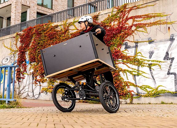 Person mit Helm fährt ein dreirädriges Lastenrad mit großer Transportbox vor einer bewachsenen Wand.