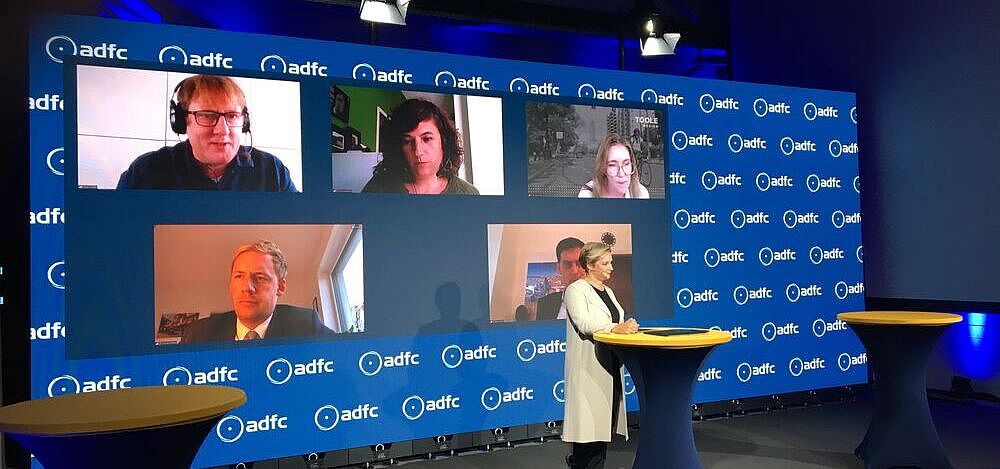 Ein Studio mit der Moderatorin am Pult, dahinter eine Videowand mit den Köpfen von per Video zugeschalteten Personen. 