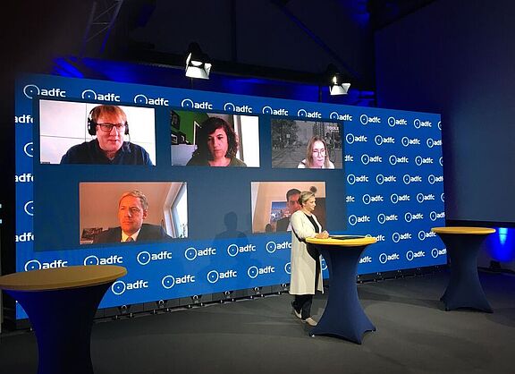 Ein Studio mit der Moderatorin am Pult, dahinter eine Videowand mit den Köpfen von per Video zugeschalteten Personen. 