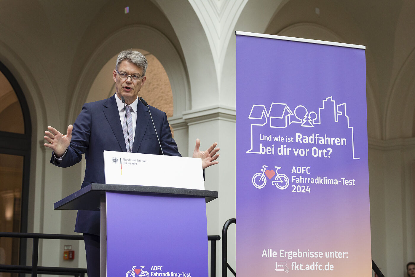 Mann mit grauen Haaren und Brille in dunklem Anzug gestikuliert am Rednerpult des Bundesverkehrsministeriums vor lila ADFC-Banner zum Fahrradklima-Test 2024