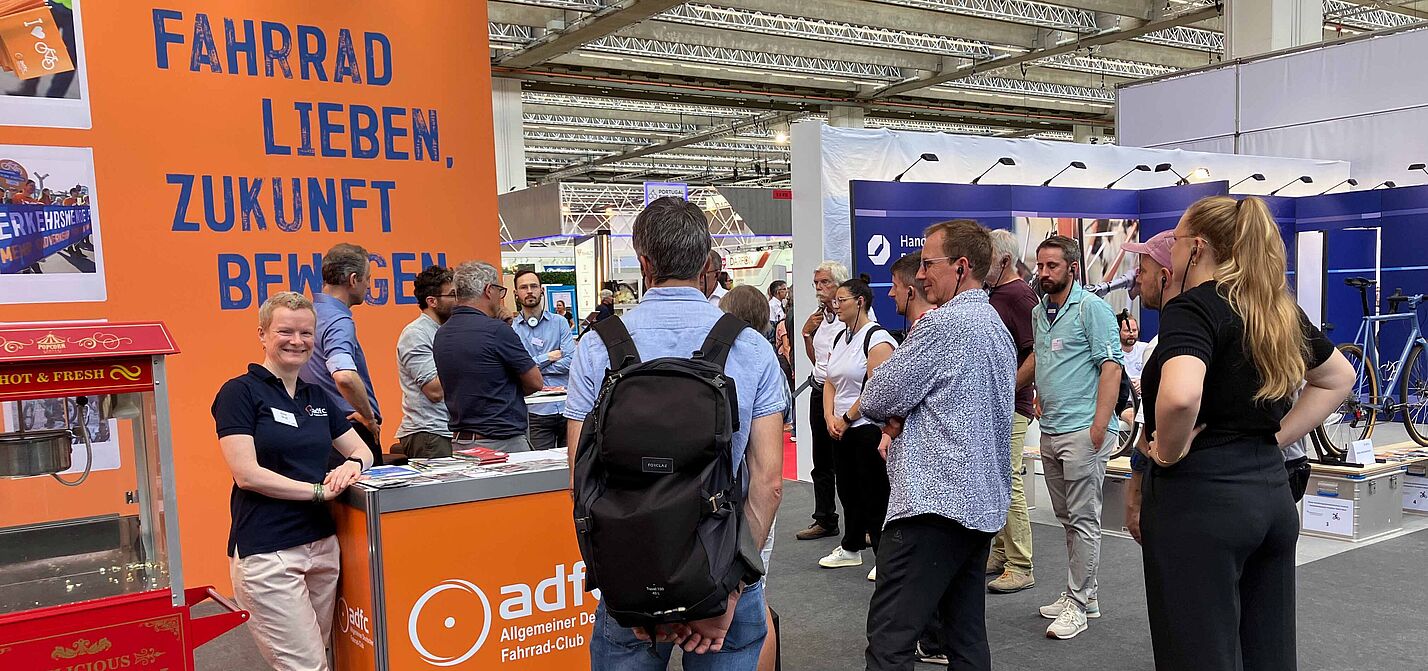 Eurobike 2025 Ein Messestand des ADFC in einer großen Halle. Vor einer orangen Rückwand mit dem blauen Slogan „Fahrrad lieben, Zukunft bewegen“ steht eine lächelnde Frau im dunkelblauen ADFC-Shirt an einer Theke. Mehrere Menschen stehen davor und dahinter in Gruppen zusammen und unterhalten sich. Im Vordergrund links ist eine rote Popcornmaschine zu sehen.
