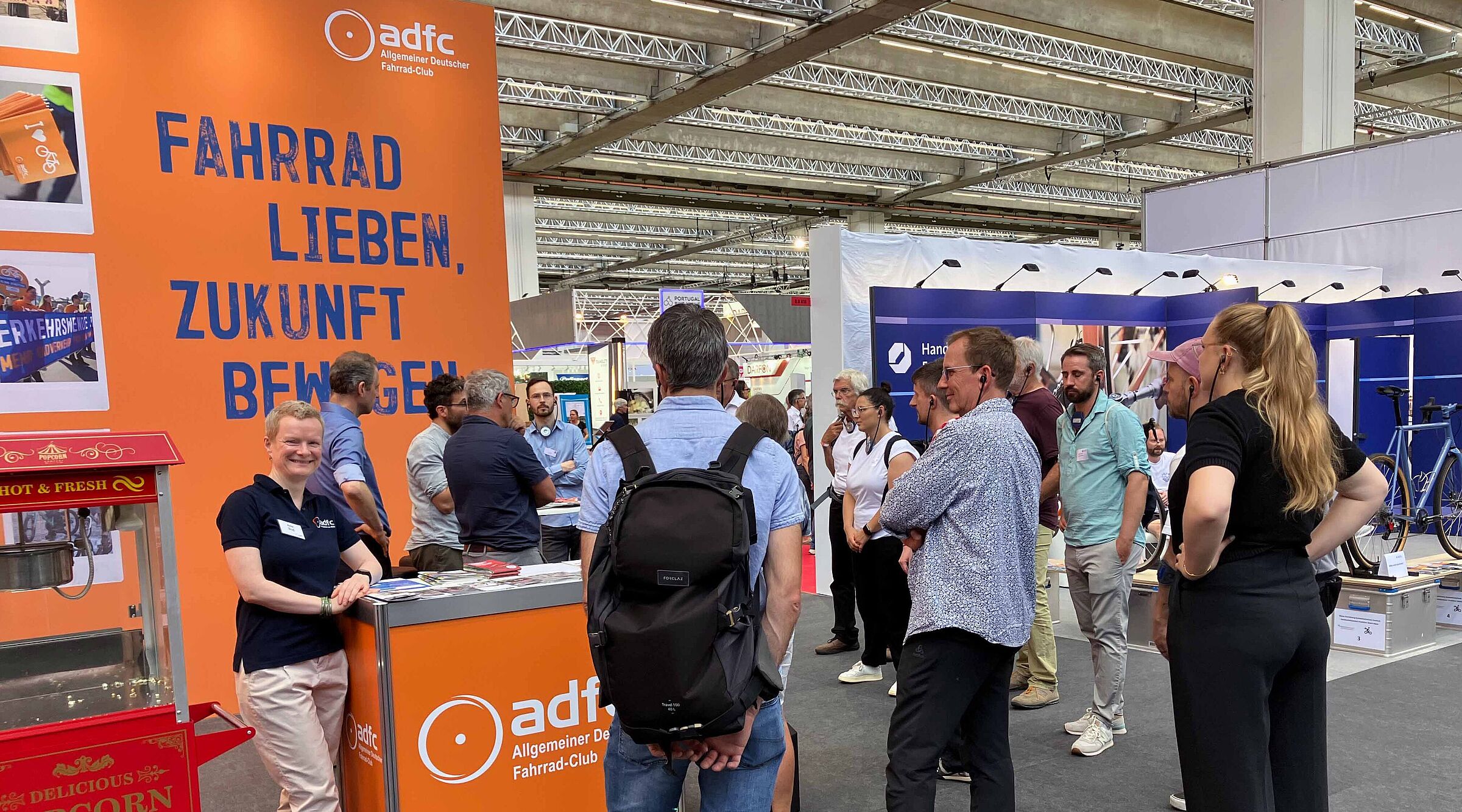 Ein Messestand des ADFC in einer großen Halle. Vor einer orangen Rückwand mit dem blauen Slogan „Fahrrad lieben, Zukunft bewegen“ steht eine lächelnde Frau im dunkelblauen ADFC-Shirt an einer Theke. Mehrere Menschen stehen davor und dahinter in Gruppen zusammen und unterhalten sich. Im Vordergrund links ist eine rote Popcornmaschine zu sehen.